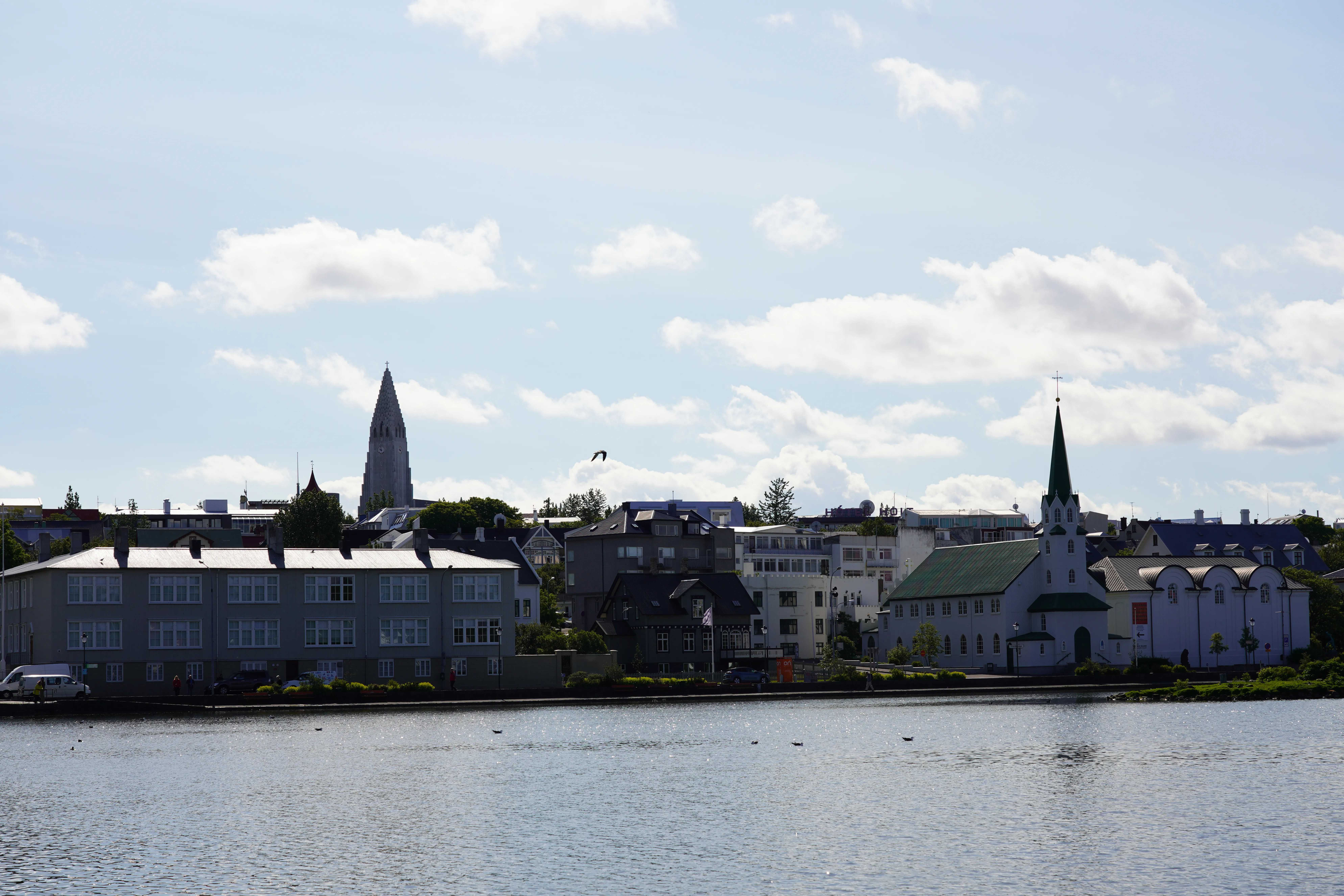 Vista de Fríkirkjan í Reykjavík con su techo verde y aguja al otro lado del lago Tjörnin, con Hallgrímskirkja visible al fondo bajo un cielo parcialmente nublado