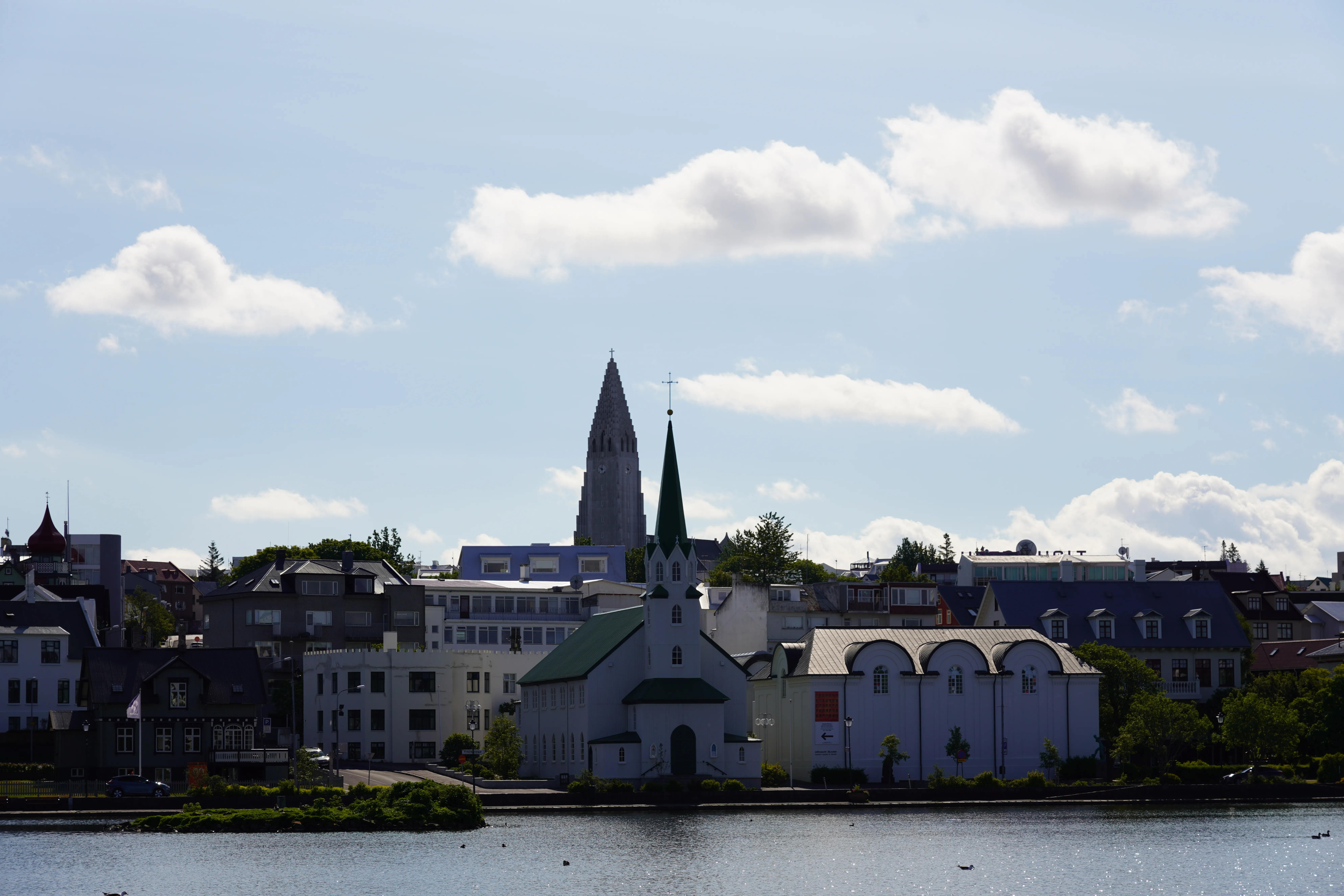 La Fríkirkjan í Reykjavík, con su tejado verde y su aguja oscura, se alza a orillas del Tjörnin, con la torre de la Hallgrímskirkja visible al fondo bajo un cielo azul parcialmente nublado.
