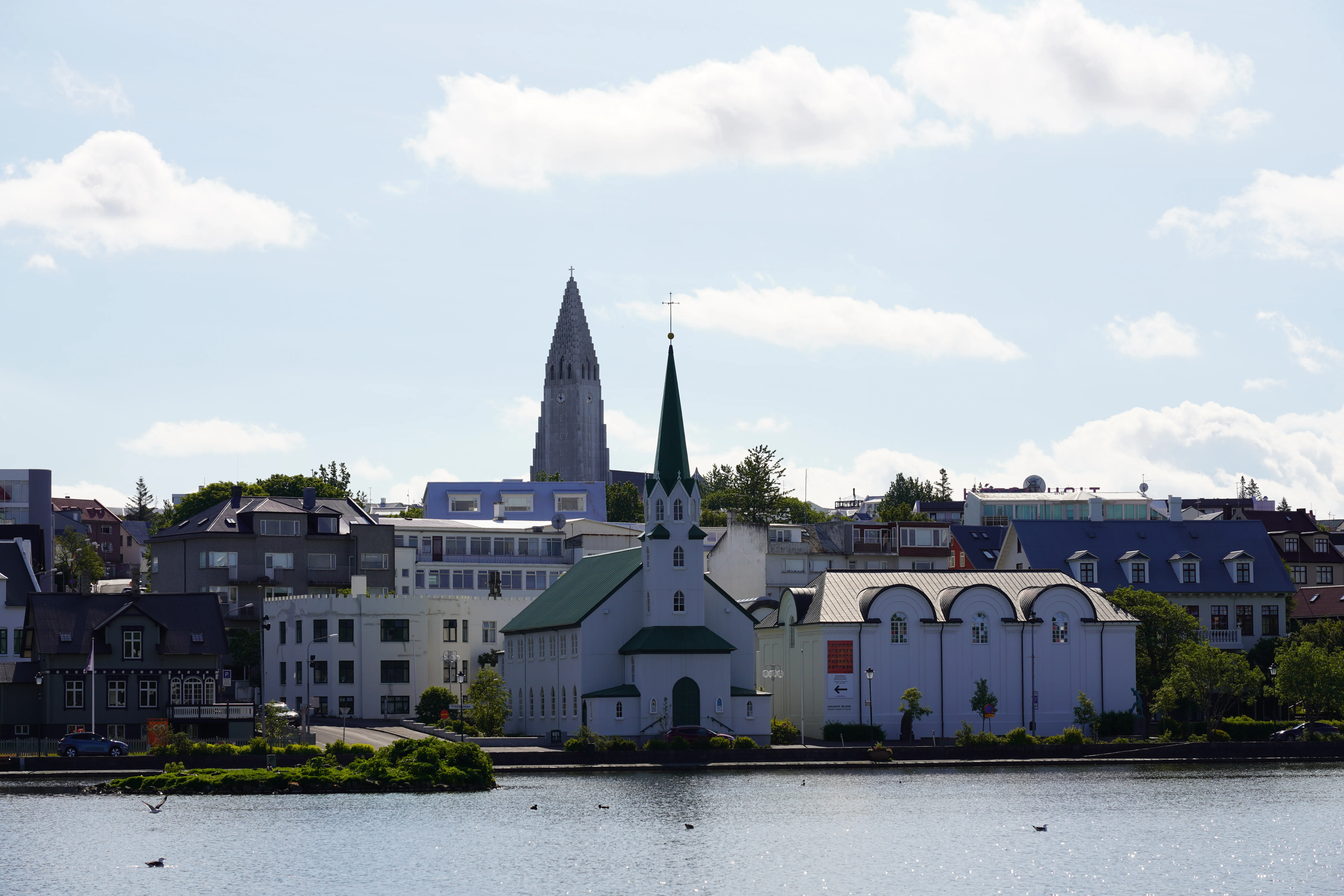 Vista exterior de la Fríkirkjan í Reykjavík, una iglesia blanca con techo y aguja verdes, situada a orillas del lago Tjörnin con la torre de la Hallgrímskirkja visible al fondo