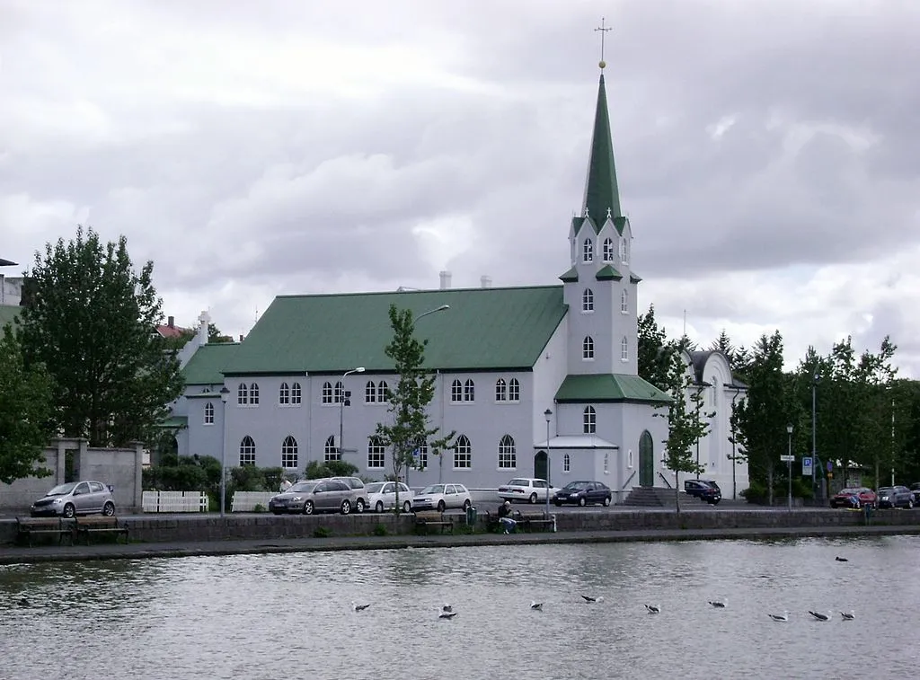 Iglesia blanca con techo verde y alta aguja verde vista desde el otro lado de un lago, con aves en el agua y coches estacionados a lo largo de la orilla bajo un cielo nublado