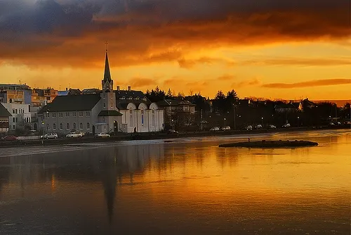 La iglesia Fríkirkjan í Reykjavík con su alta aguja reflejada en las aguas del lago Tjörnin bajo un espectacular cielo de atardecer naranja.