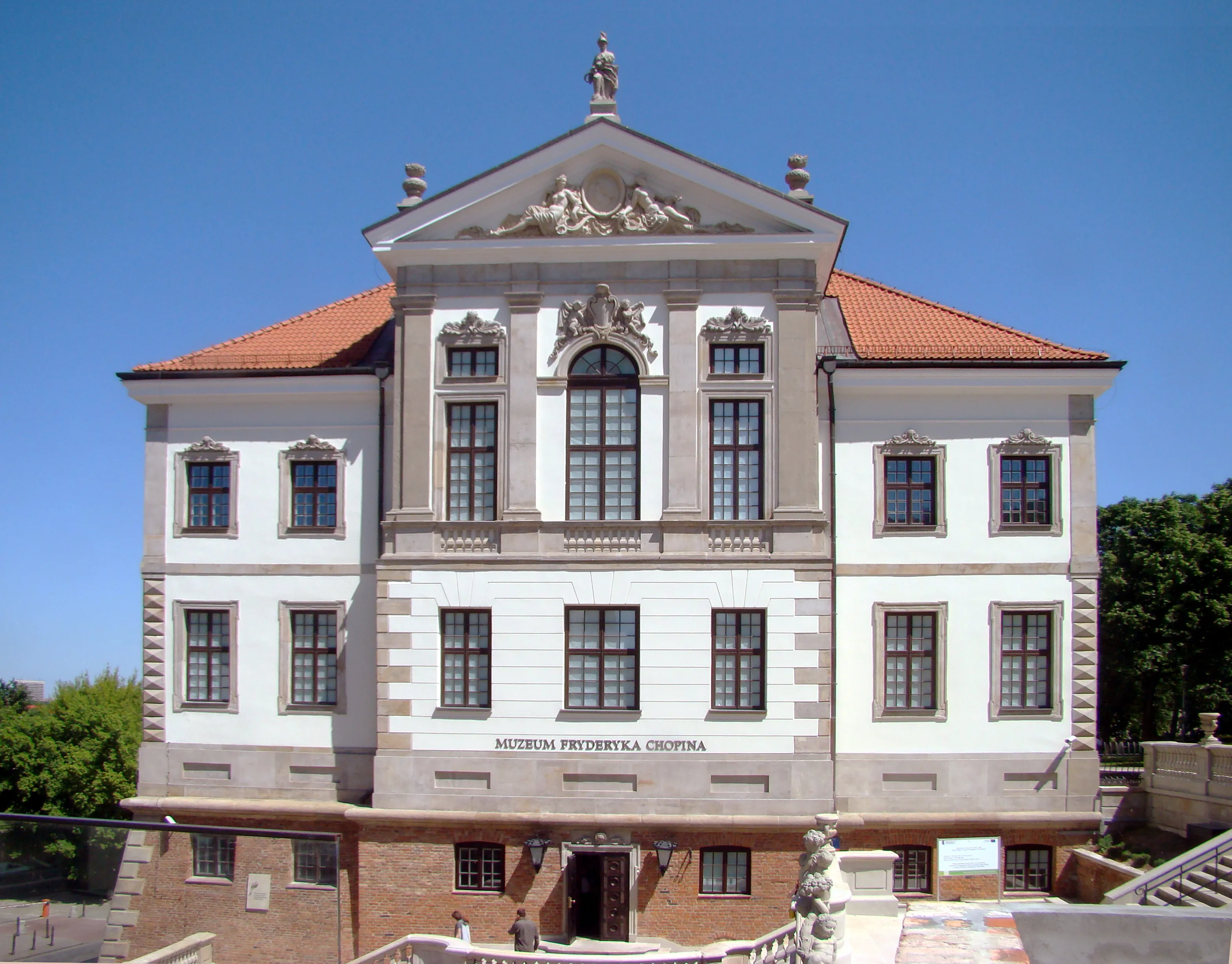 The Fryderyk Chopin Museum building with a white facade, red roof, and a sign reading 'MUZEUM FRYDERYKA CHOPINA' under a clear blue sky