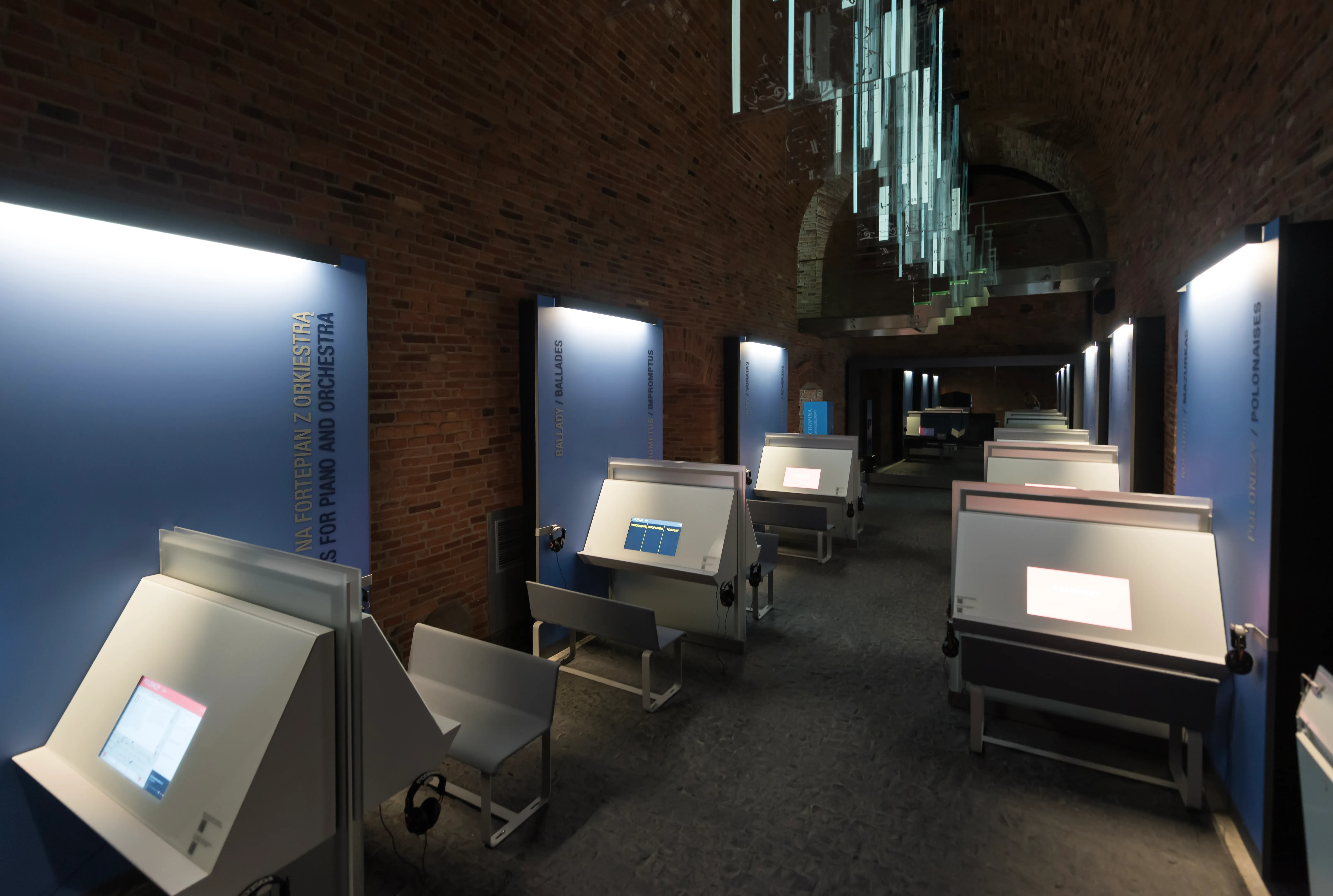 Interior hallway with interactive display stations, blue-lit panels, and a chandelier in a brick-walled gallery