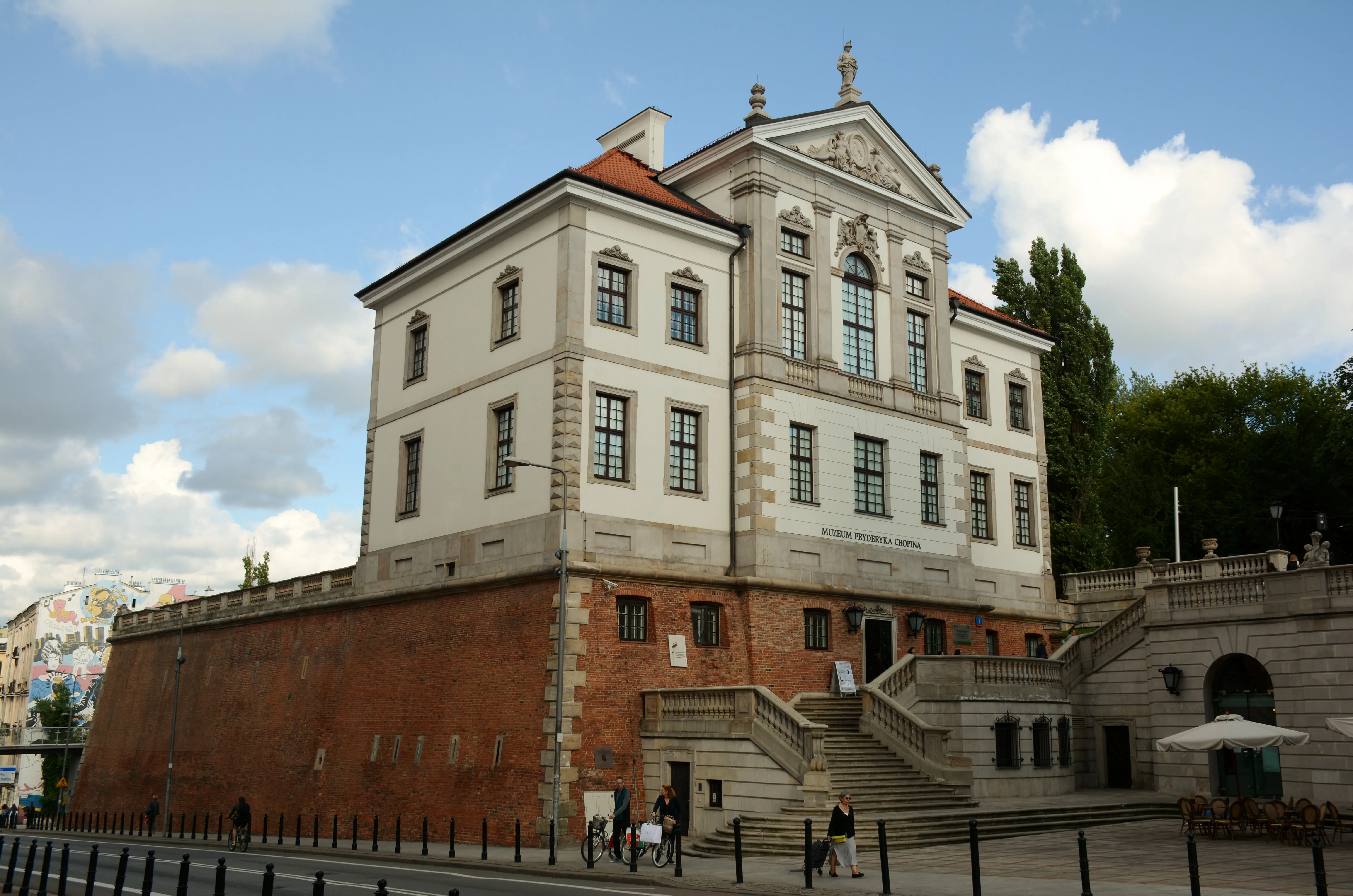 Neoclassical building with white upper floors and red brick lower level, featuring large windows, a central staircase, and a decorative pediment under a blue sky.