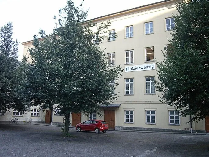 Buitenaanzicht van het gebouw FÜNFZIGZWANZIG met een bord met het opschrift 'fünfzigzwanzig' op de beige gevel, grote bomen op de binnenplaats en een rode auto geparkeerd aan de voorzijde.