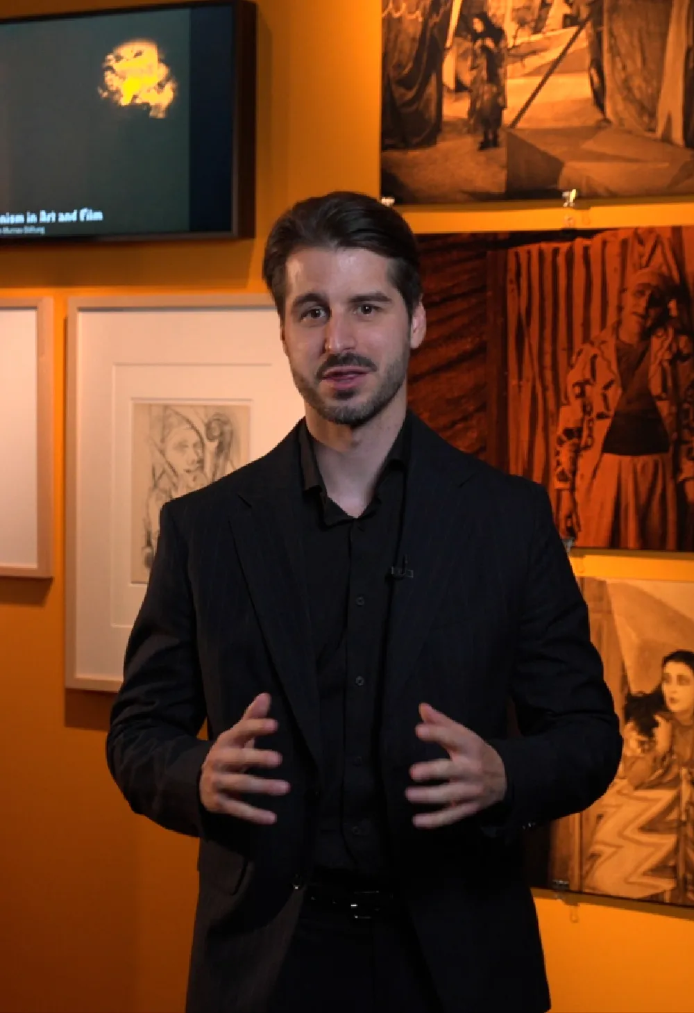 Man in een zwart pak, staand in een kunstgalerie met ingelijste kunstwerken en een scherm op de achtergrond