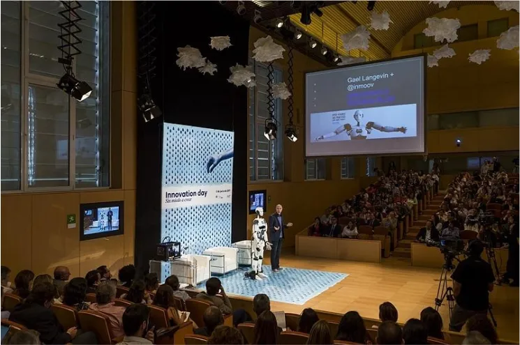 Un gran auditorio con un escenario, un orador, una pantalla que muestra una presentación y una audiencia sentada en filas.