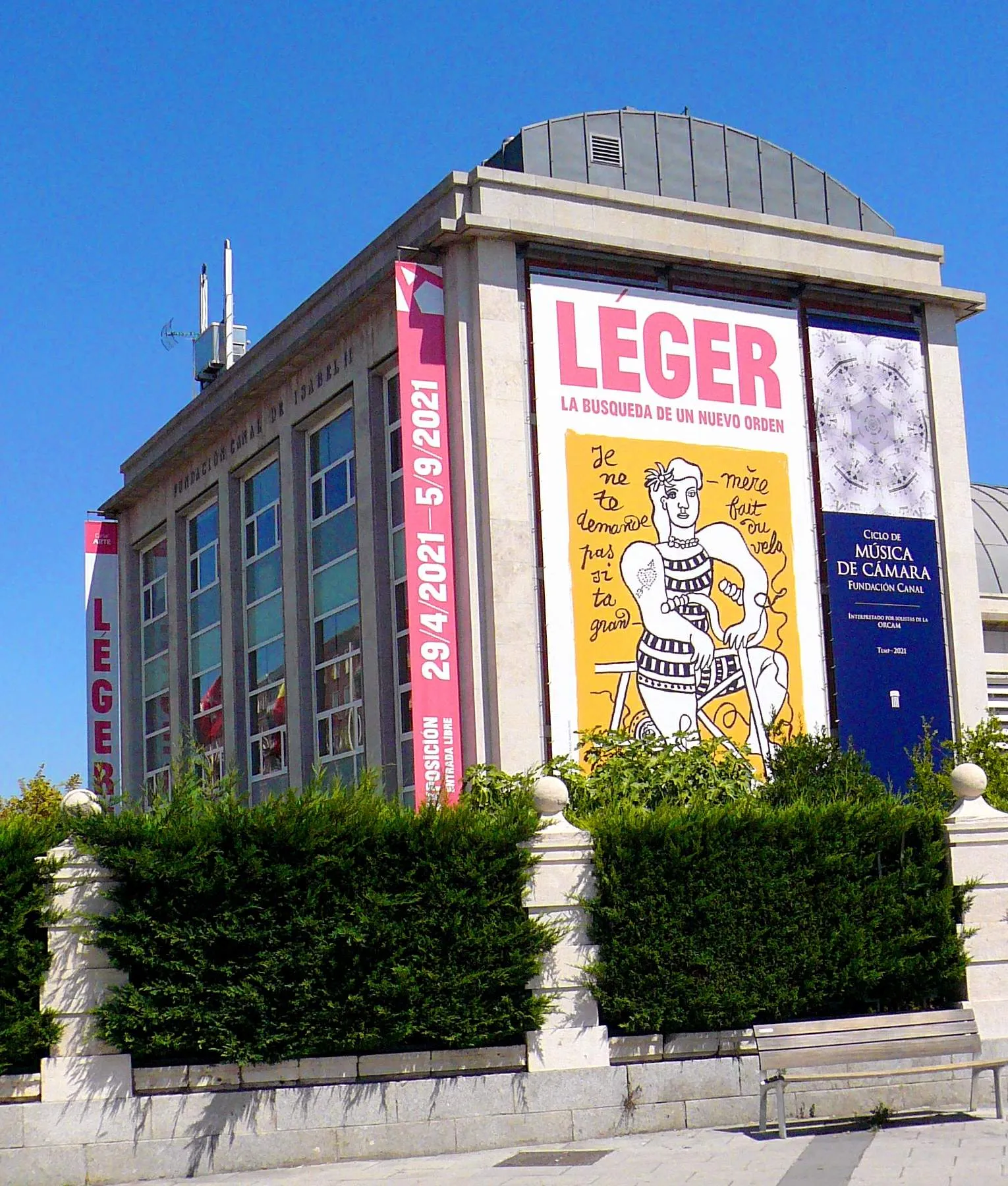 Exterior del edificio con grandes pancartas de exposiciones de arte, setos verdes delante y un cielo azul claro.