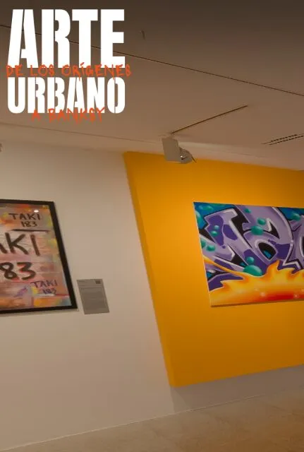 Een galeriewand met een grote kleurrijke graffiti schildering op een geel paneel en een ingelijst kunstwerk met tekst op een witte muur.