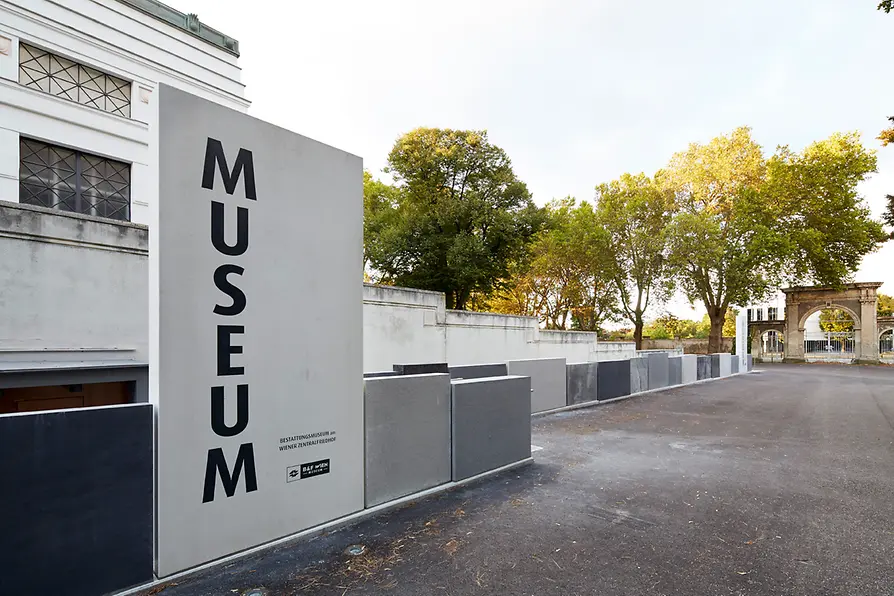 Een modern gebouw met een groot wit bord met de tekst MUSEUM in zwarte letters, tegen een achtergrond van bomen en een verhard pad op de Wiener Zentralfriedhof.