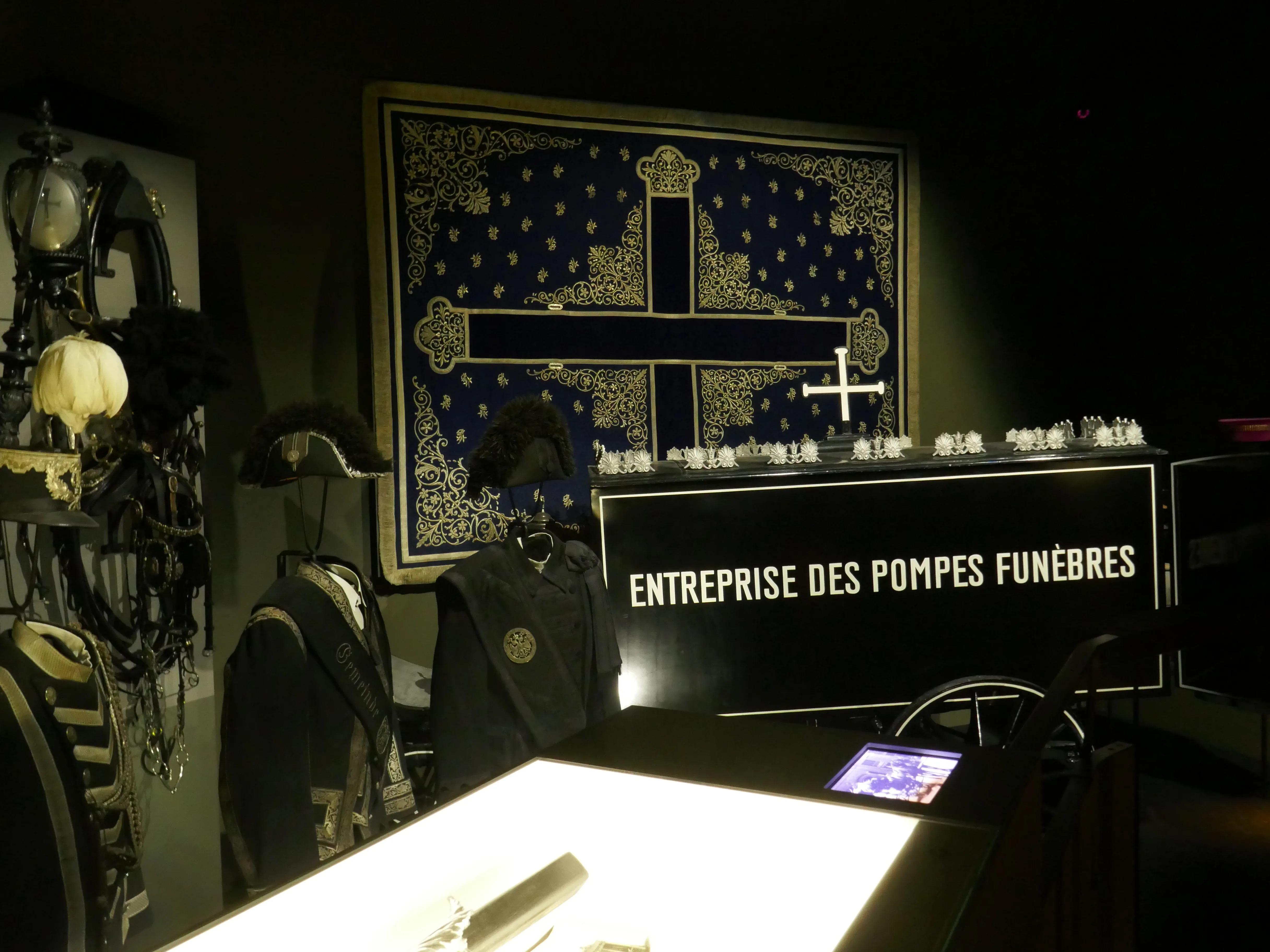 Interieurtentoonstelling met een bord waarop 'ENTREPRISE DES POMPES FUNEBRES' staat, een decoratief wandkleed met een kruis en tentoongestelde parafernalia gerelateerd aan begrafenissen.