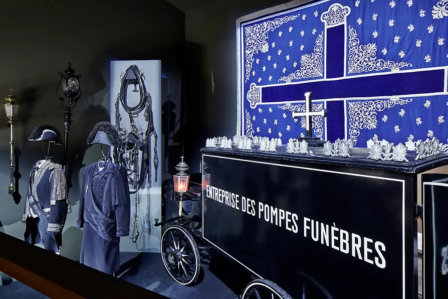 Tentoonstelling met een zwarte lijkwagen, paspoppen in rouwkleding en een blauw-wit spandoek met een kruis