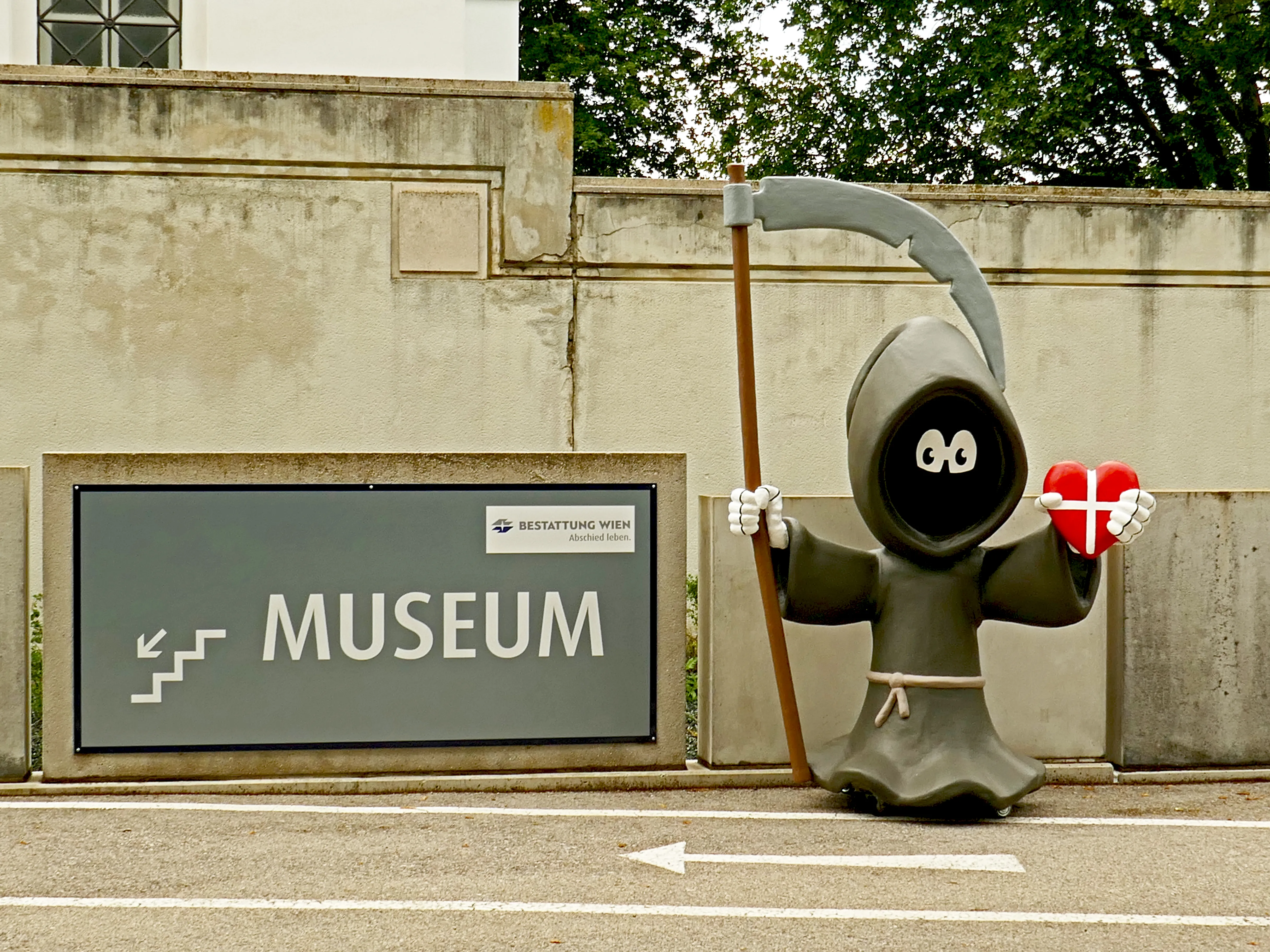 Een grijs standbeeld van een Magere Hein die een rood hart en een zeis vasthoudt, staat naast een bord met de tekst 'MUSEUM' op een betonnen muur.