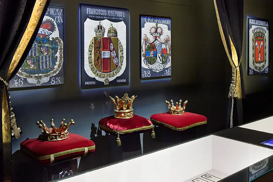 Drie kronen op rode kussens tentoongesteld in een vitrine met historische wapenschildposters aan de muur