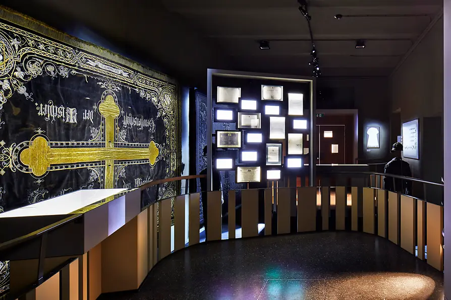 Interieur van een museum met een groot zwart wandkleed met een gouden kruis en tekst, naast een muur met verlichte vitrines.