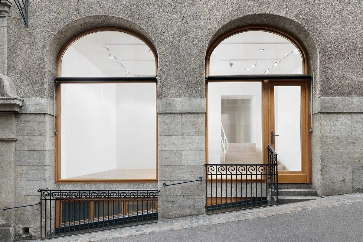 Fachada de piedra con dos grandes ventanas arqueadas y una puerta de entrada de madera, con barandillas de hierro negro a lo largo de una calle empedrada.