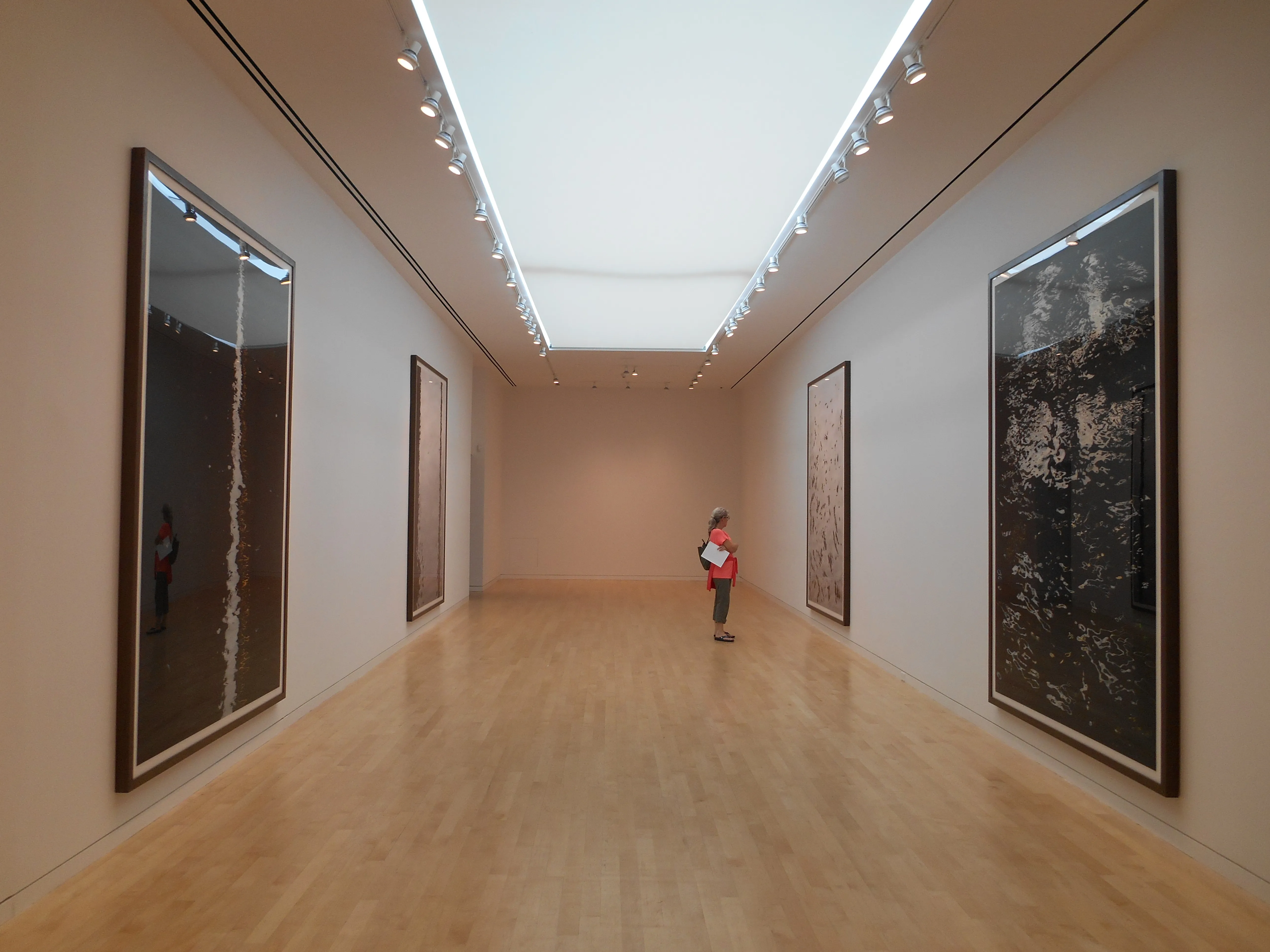 Largo pasillo de galería con grandes obras de arte enmarcadas en ambas paredes, un techo con tragaluz luminoso y suelos de madera clara, con un visitante de pie al fondo.
