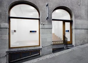 Fachada de piedra gris con dos grandes ventanales arqueados que muestran el interior de la galería, una barandilla de metal negro a lo largo de la acera y una entrada con escaleras visible.