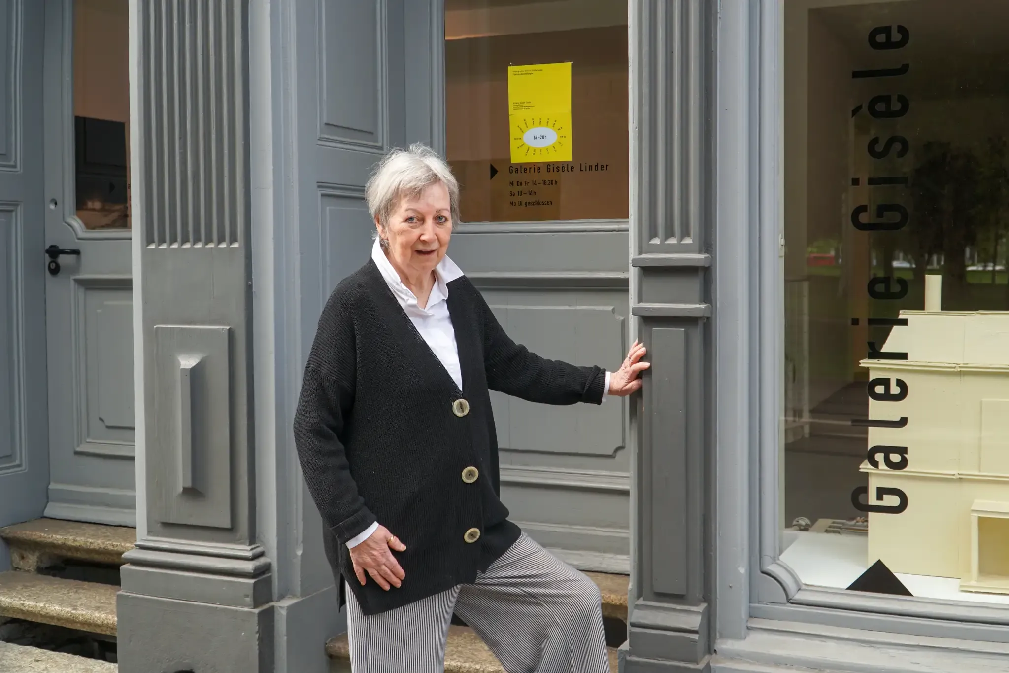 Mujer con cárdigan negro de pie en la entrada gris de un edificio, con el texto Galerie Gisèle visible en la ventana adyacente