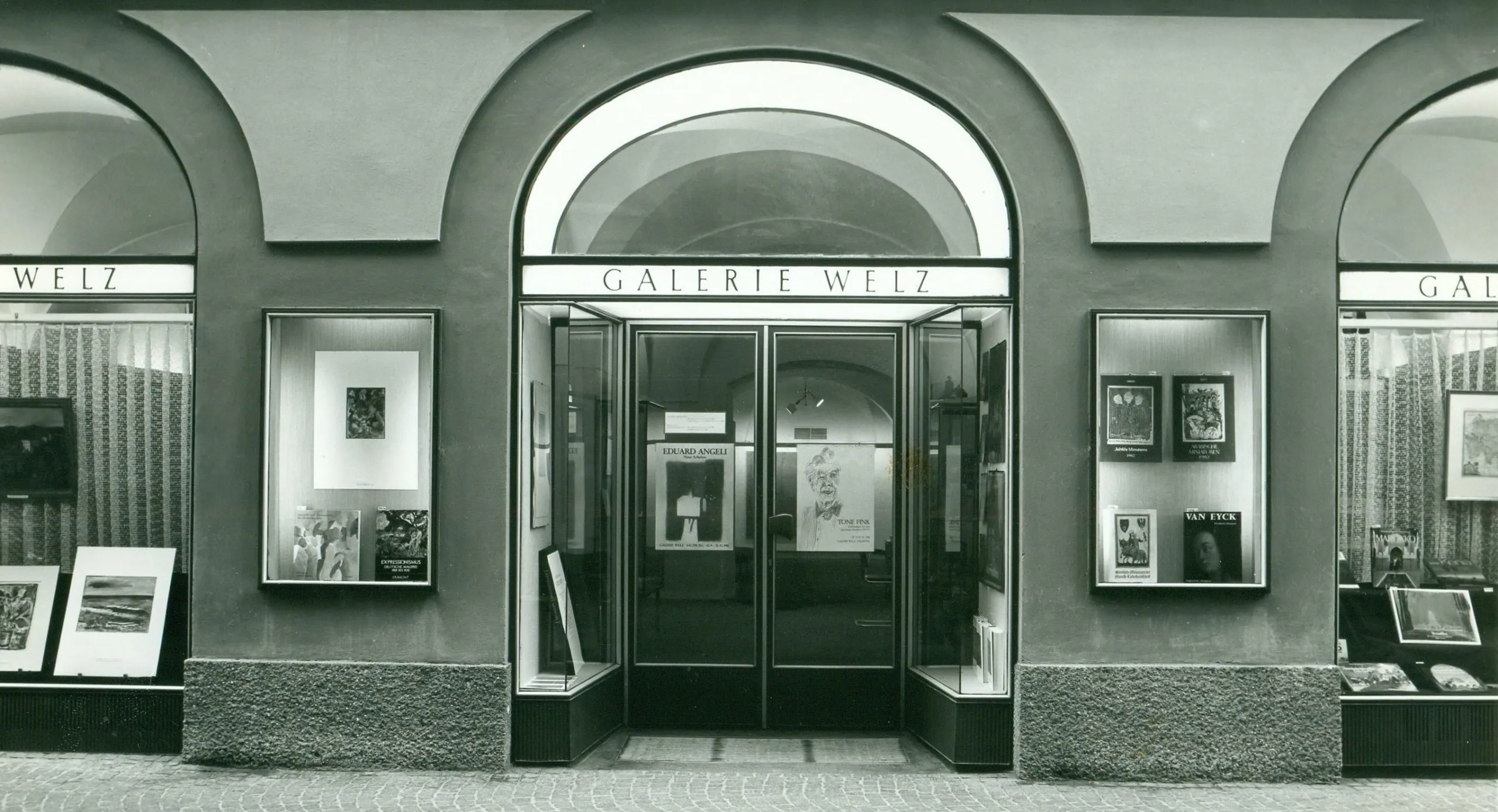 Vista exterior en blanco y negro de la fachada de la Galerie Welz, que cuenta con ventanas arqueadas y vitrinas con obras de arte enmarcadas.