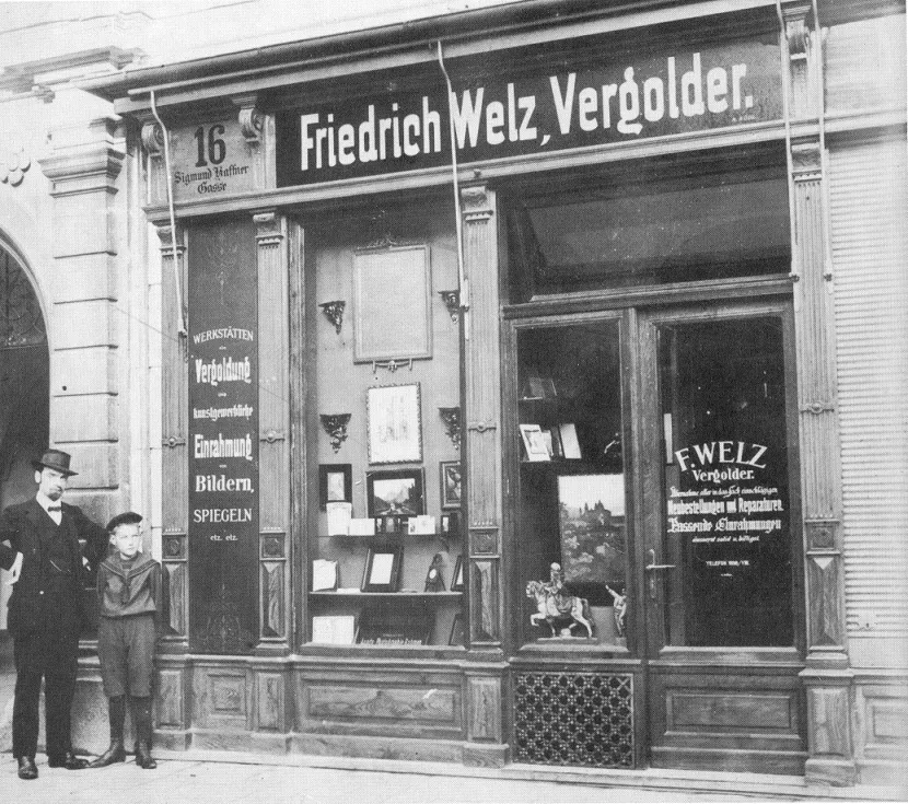 Fotografía histórica en blanco y negro del escaparate de Friedrich Welz, Vergolder, con dos personas de pie frente a la entrada.