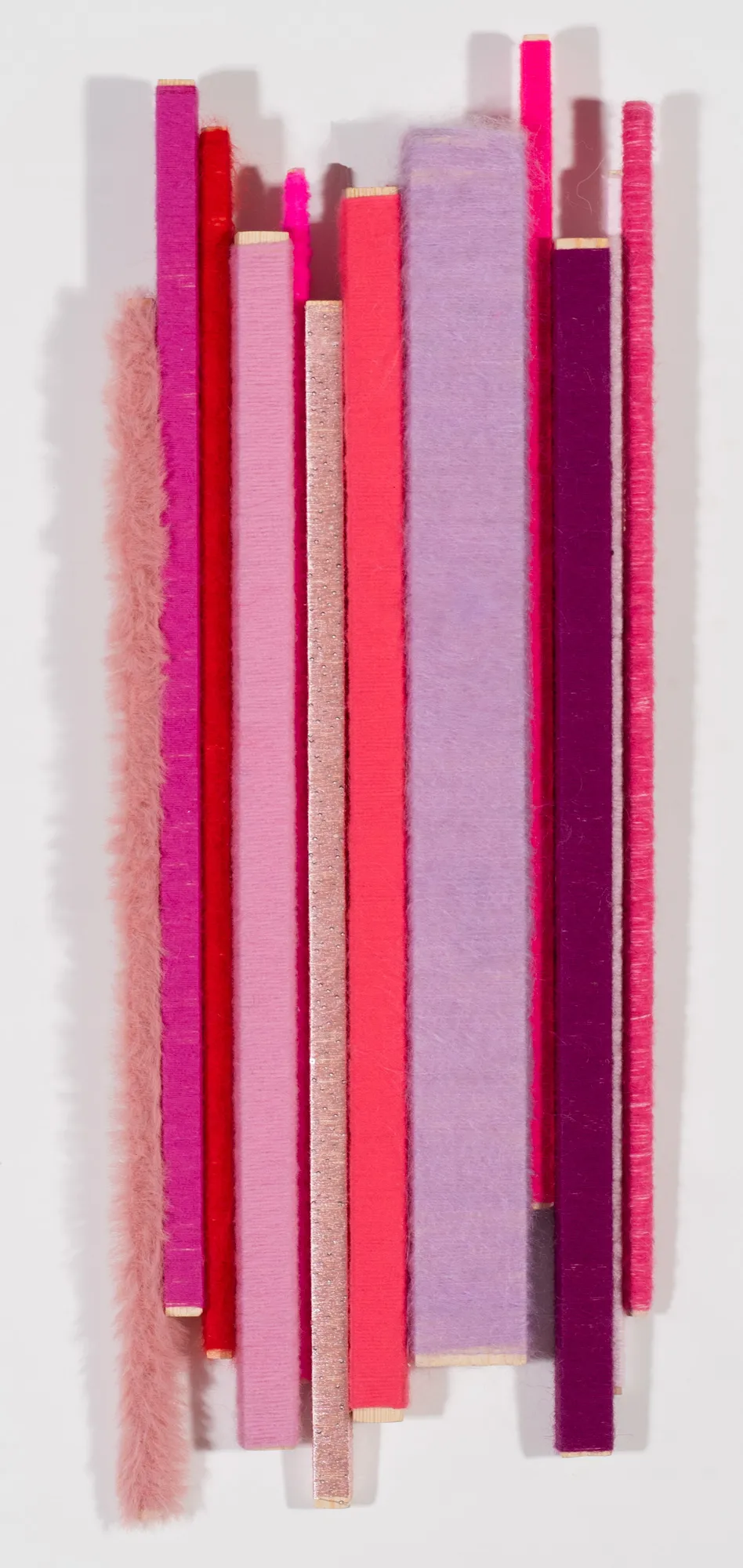 Tiras verticales de materiales texturizados en tonos rosa, magenta, rojo y lavanda dispuestas sobre un fondo blanco, proyectando sombras suaves