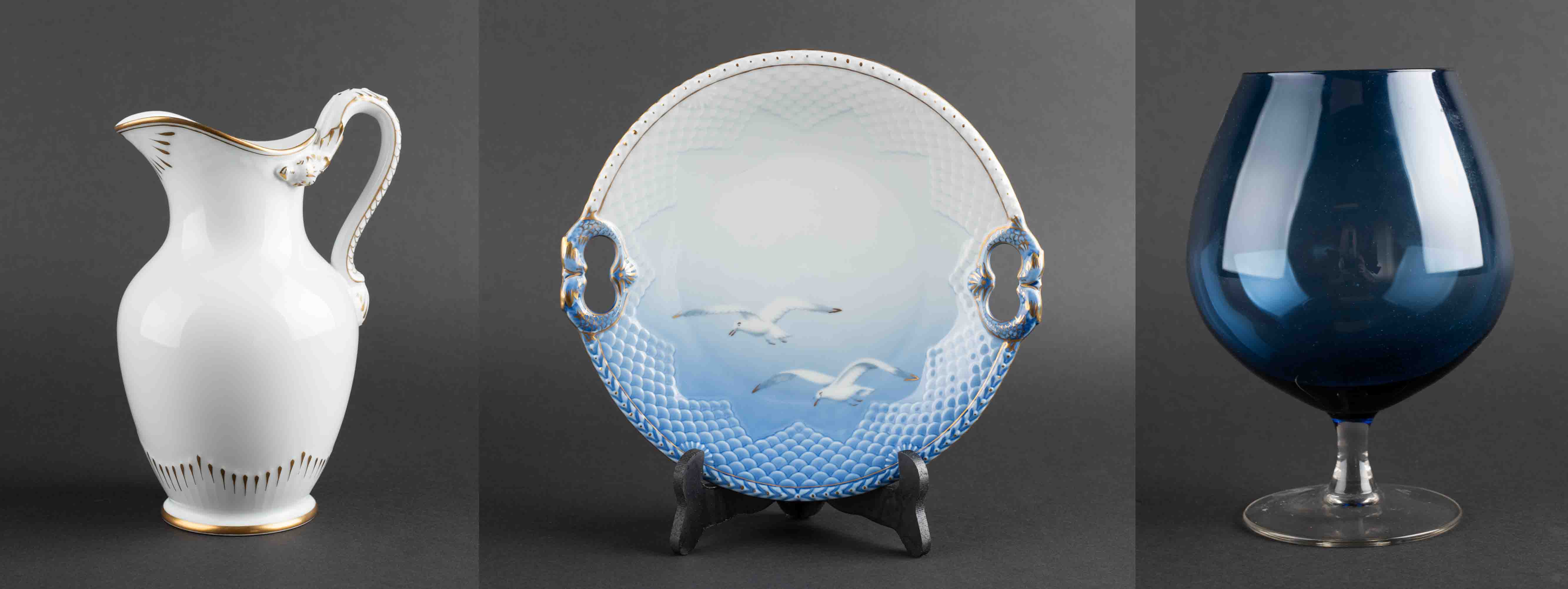 Tres objetos sobre un fondo oscuro: una jarra de cerámica blanca, un plato azul y blanco con gaviotas sobre un soporte y una copa de cristal azul oscuro.