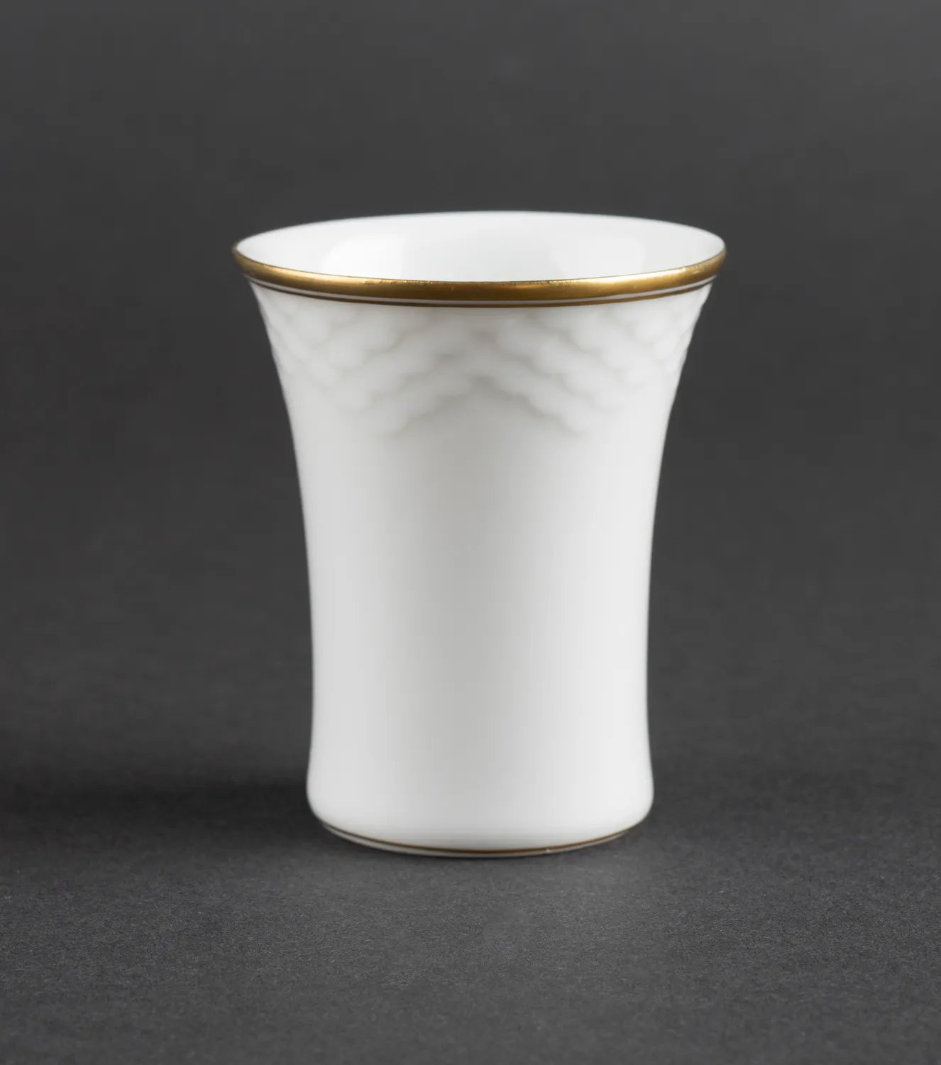 Taza de cerámica blanca con borde acampanado y borde dorado fotografiada sobre un fondo gris oscuro