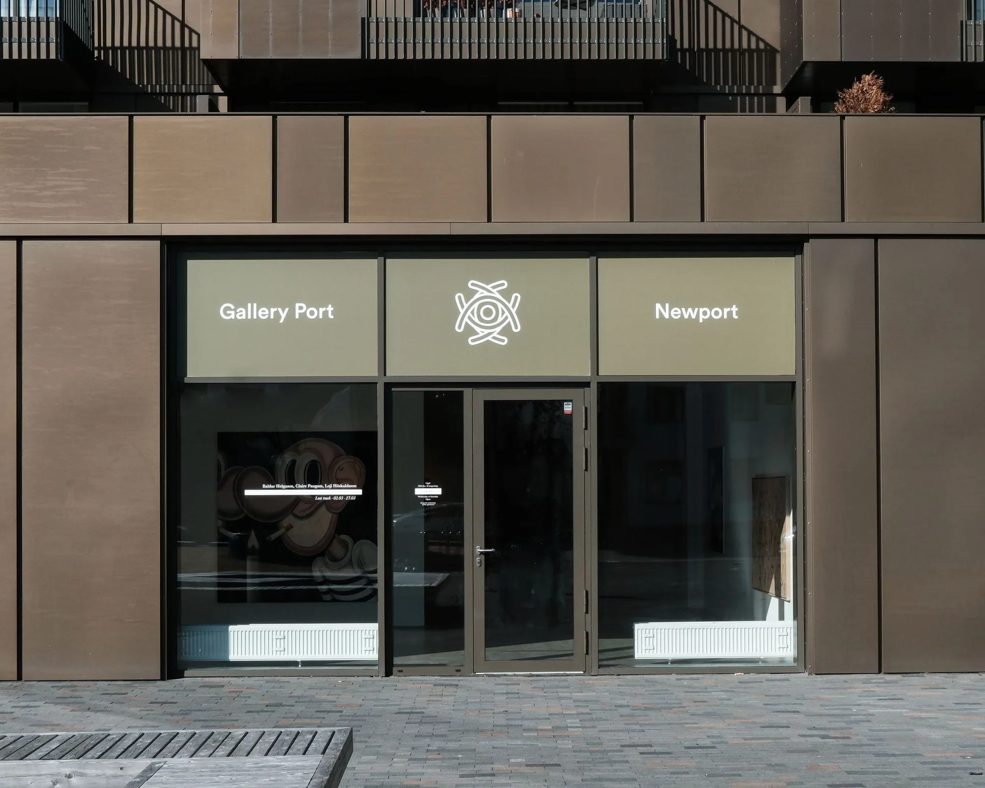 Escaparate a nivel de calle de Gallery Port con fachada de paneles marrones, puertas de entrada de cristal y rótulos con los nombres Gallery Port y Newport flanqueando un logotipo central.