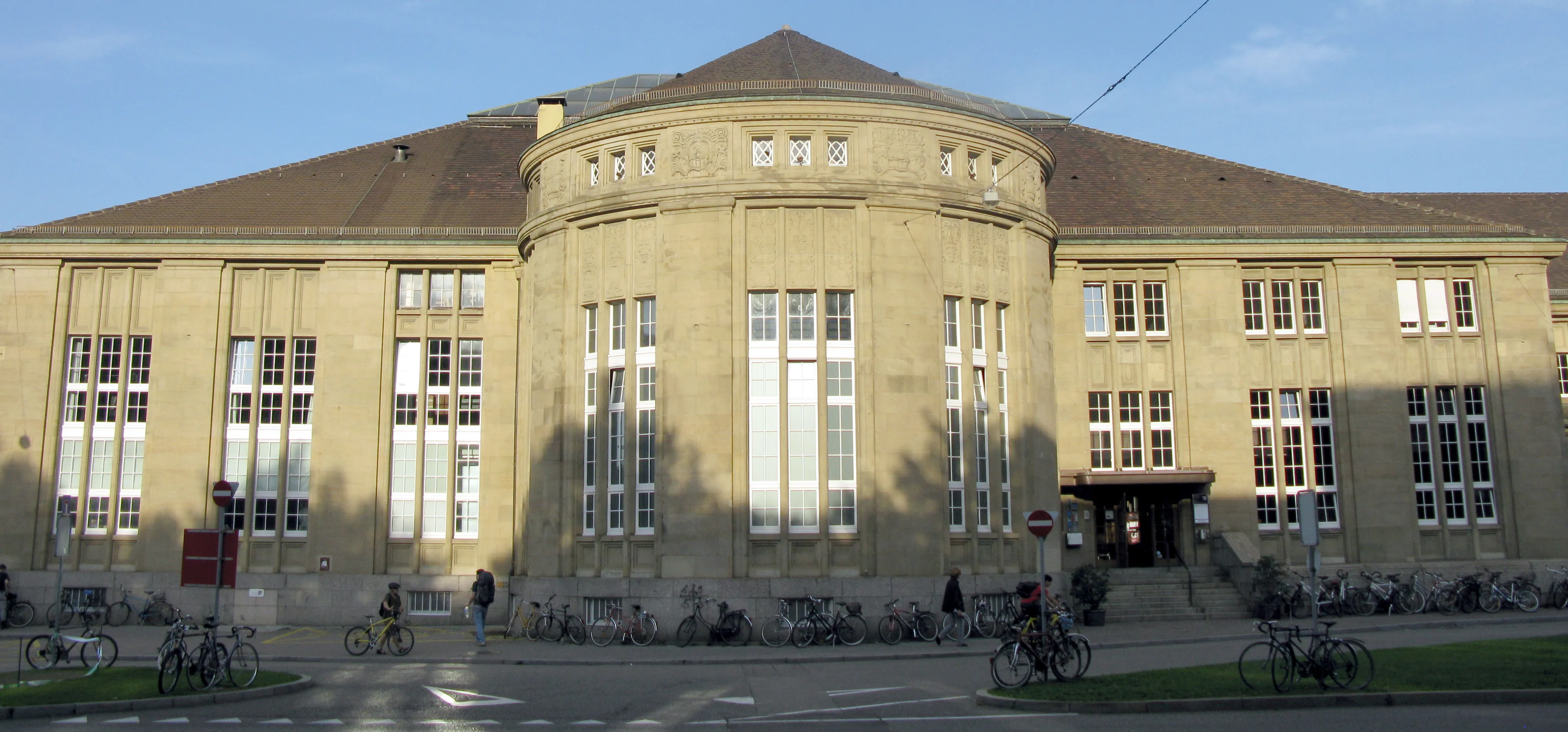 Szeroki widok na budynek Gare du Nord w Bazylei, dużą konstrukcję z beżowego kamienia z zaokrągloną środkową częścią fasady, wysokimi pionowymi oknami i licznymi rowerami zaparkowanymi wzdłuż frontu.