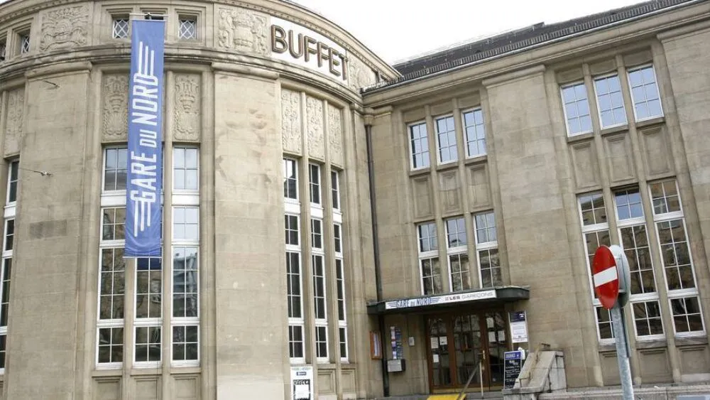 Zewnętrzna fasada kamiennego budynku z niebieskim pionowym banerem z napisem 'GARE du NORD' oraz słowem 'BUFFET' wyrytym nad oknami.