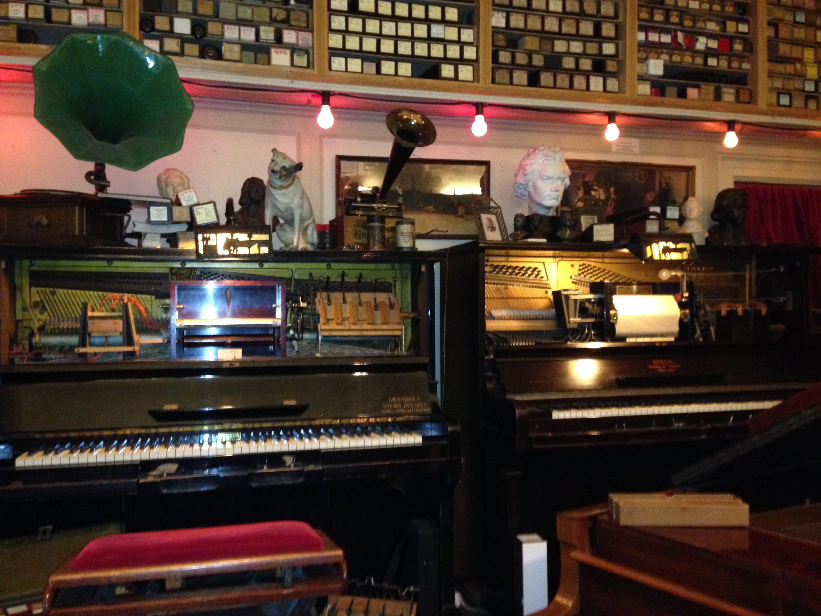 Several antique pianos and mechanical music instruments on display in a warmly lit museum room with shelves of music rolls.