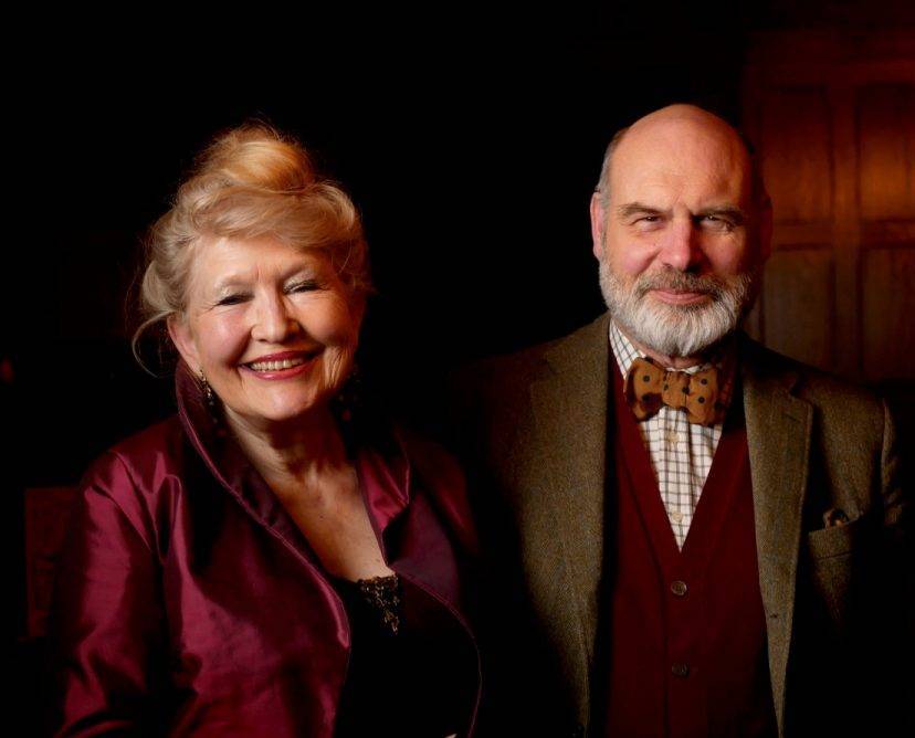 Two people posing together, woman in burgundy jacket and man in brown suit with red vest and bow tie, dark background