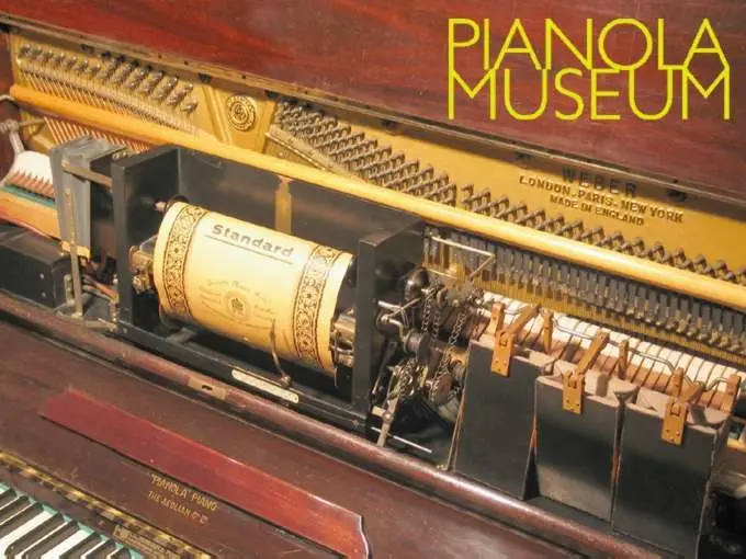 Close-up of a pianola's internal mechanism showing a paper music roll labeled 'Standard' with yellow 'PIANOLA MUSEUM' text overlay