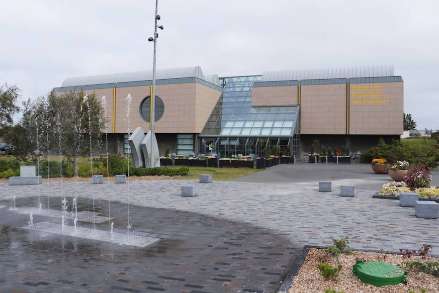 Vista exterior del Gerðarsafn Art Museum que muestra la moderna fachada del edificio con marquesina de entrada de cristal, una ventana circular y una fuente en la plaza pavimentada en primer plano.