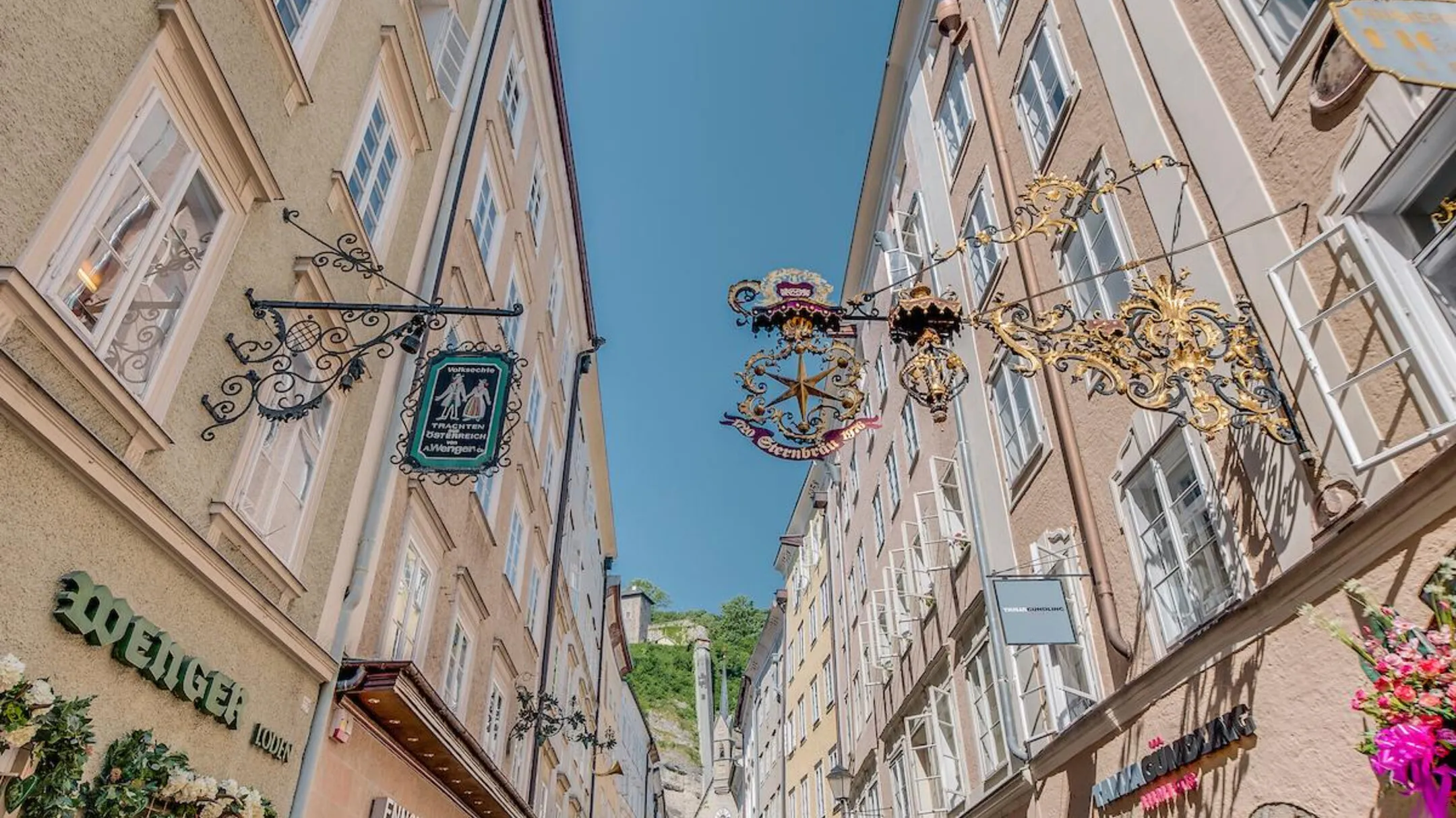 Calle peatonal estrecha con letreros gremiales de hierro forjado ornamentado que cuelgan de casas históricas de colores pastel bajo un cielo azul