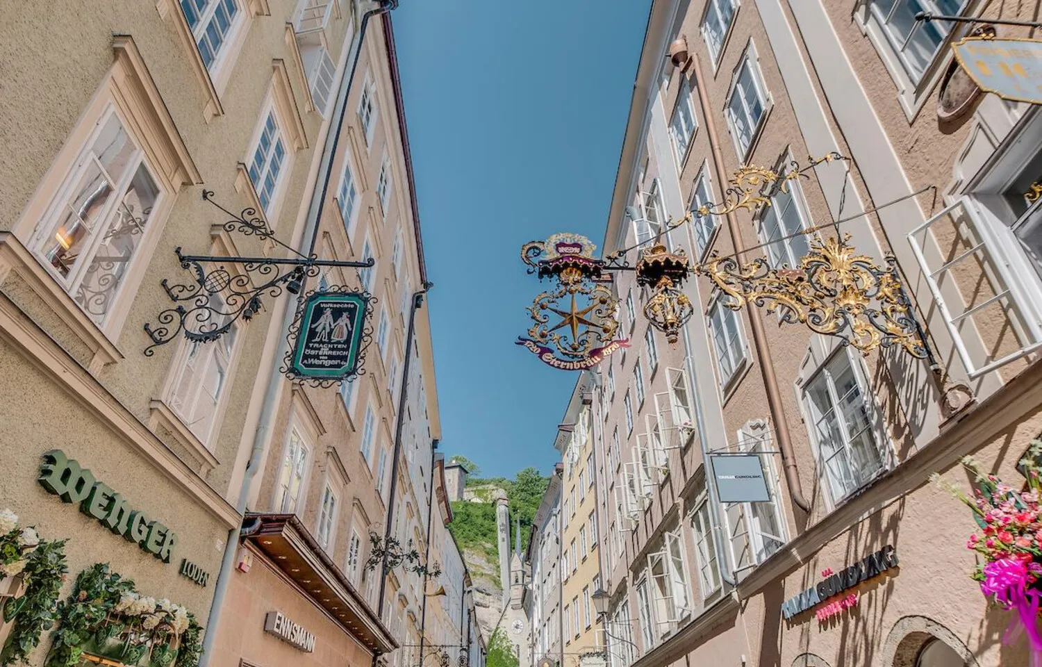 Vista en ángulo contrapicado de edificios históricos y ornamentados carteles gremiales de hierro forjado a lo largo de la estrecha calle Getreidegasse bajo un cielo azul despejado.