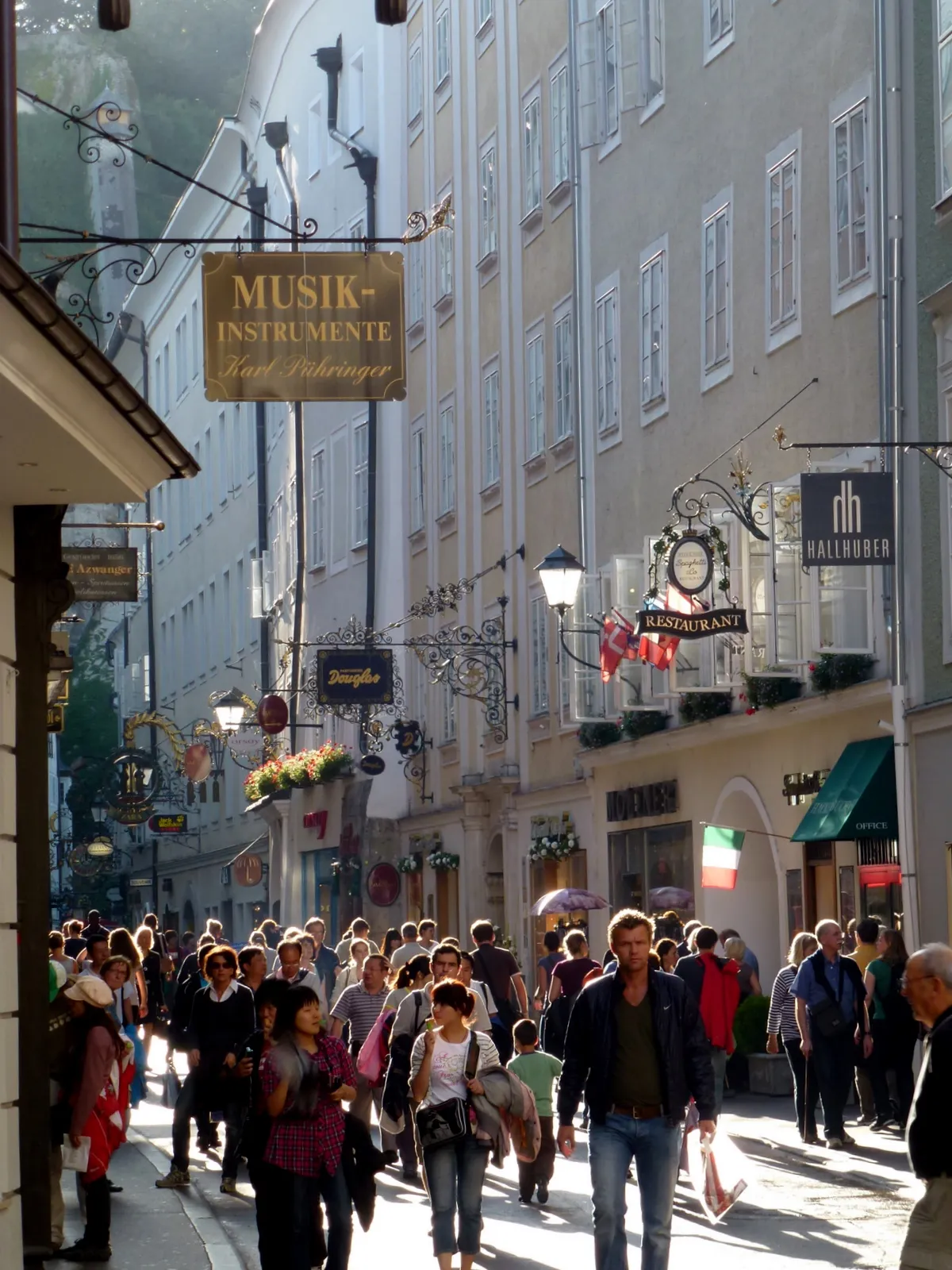 Calle peatonal concurrida con letreros gremiales de hierro forjado ornamentados, incluido un prominente cartel de Musik-Instrumente, y casas tradicionales que bordean la estrecha Getreidegasse en Salzburgo.