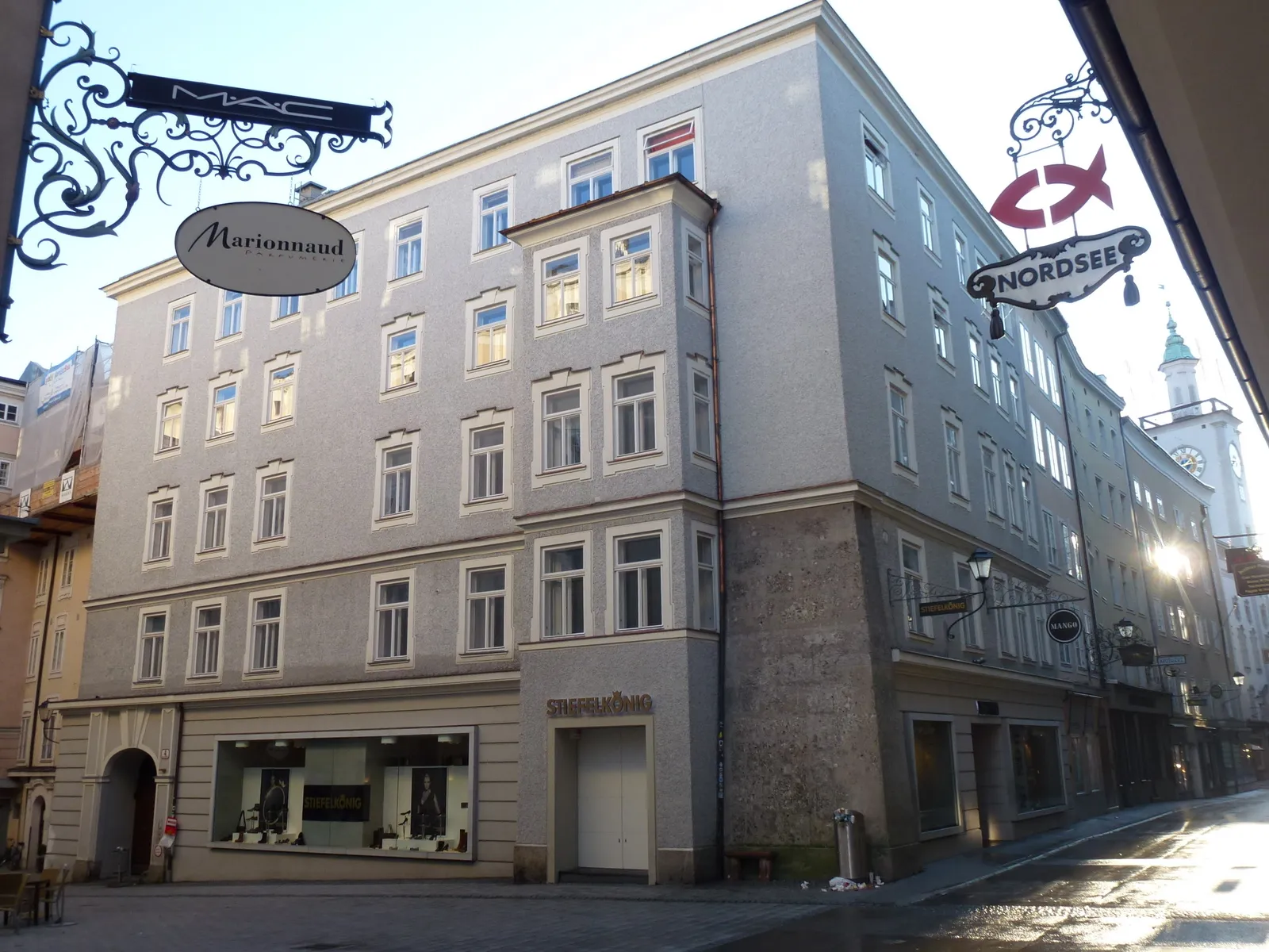 Una casa adosada de varios pisos de color gris claro en una calle estrecha que presenta letreros ornamentados de hierro forjado, incluido uno de 'Nordsee', con la aguja de una iglesia visible al fondo.