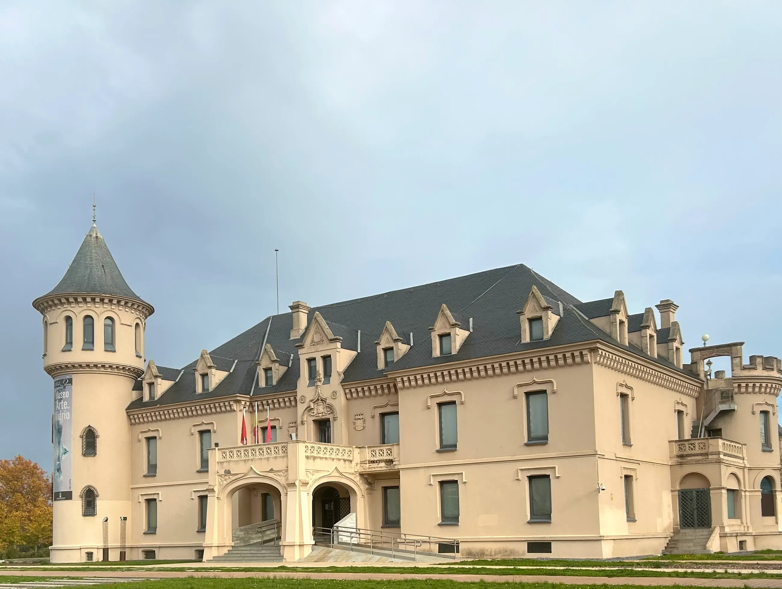 Un gran edificio beige parecido a un castillo con una torre cónica, múltiples ventanas y una entrada central bajo un cielo nublado.