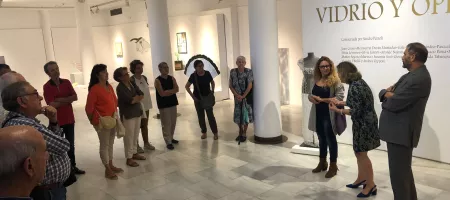 Grupo de personas viendo instalaciones de arte de vidrio en una galería de museo con paredes y columnas blancas.