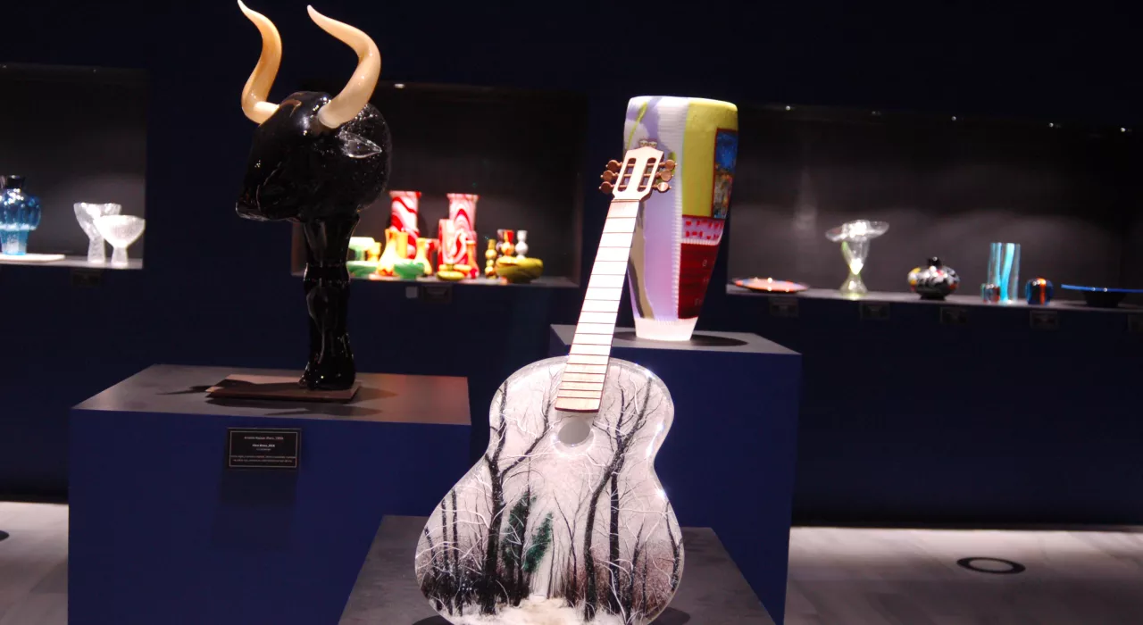 Exposición de arte en vidrio con una escultura de toro y una escultura de guitarra expuestas en una galería oscura