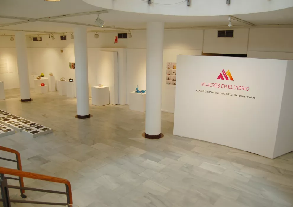 Galería interior blanca con columnas, vitrinas y un gran letrero con el logo del museo y el texto 'Mujeres en el Vidrio'