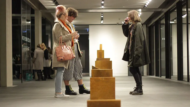 Visitantes observando una instalación de esculturas de bloques de madera apilados en una galería moderna con paredes de cristal e iluminación de riel.