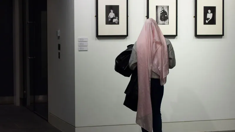Persona con una tela rosa sobre la cabeza parada en una galería mirando tres retratos fotográficos en blanco y negro enmarcados en una pared blanca