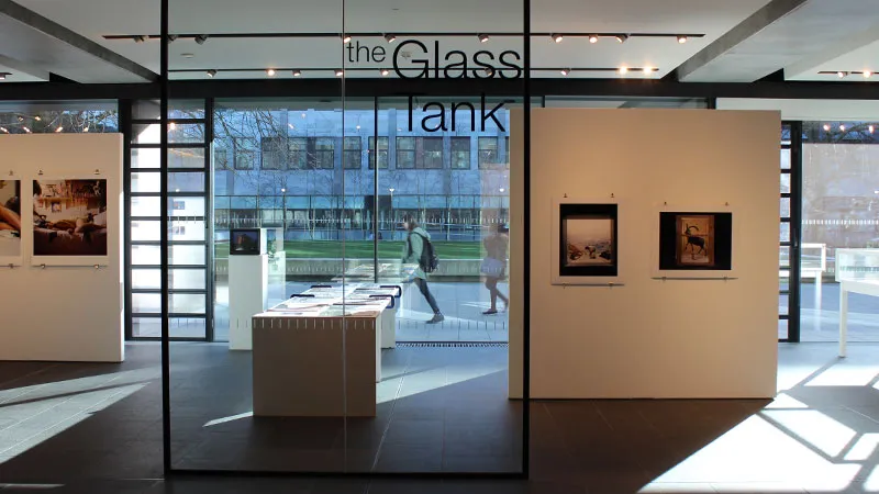 Interior del espacio de exposición The Glass Tank que muestra paredes de cristal con el nombre del lugar, paneles divisorios blancos con obras de arte enmarcadas, una vitrina central y personas visibles a través del cristal.