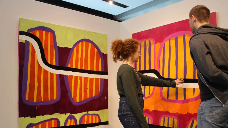 Dos personas observan grandes pinturas abstractas y coloridas con formas llamativas en naranja, morado y rosa en una galería de exposiciones