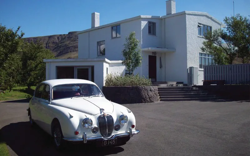 Casa blanca de dos pisos con un Jaguar blanco vintage aparcado en el camino de grava, colinas verdes visibles al fondo bajo un cielo azul despejado