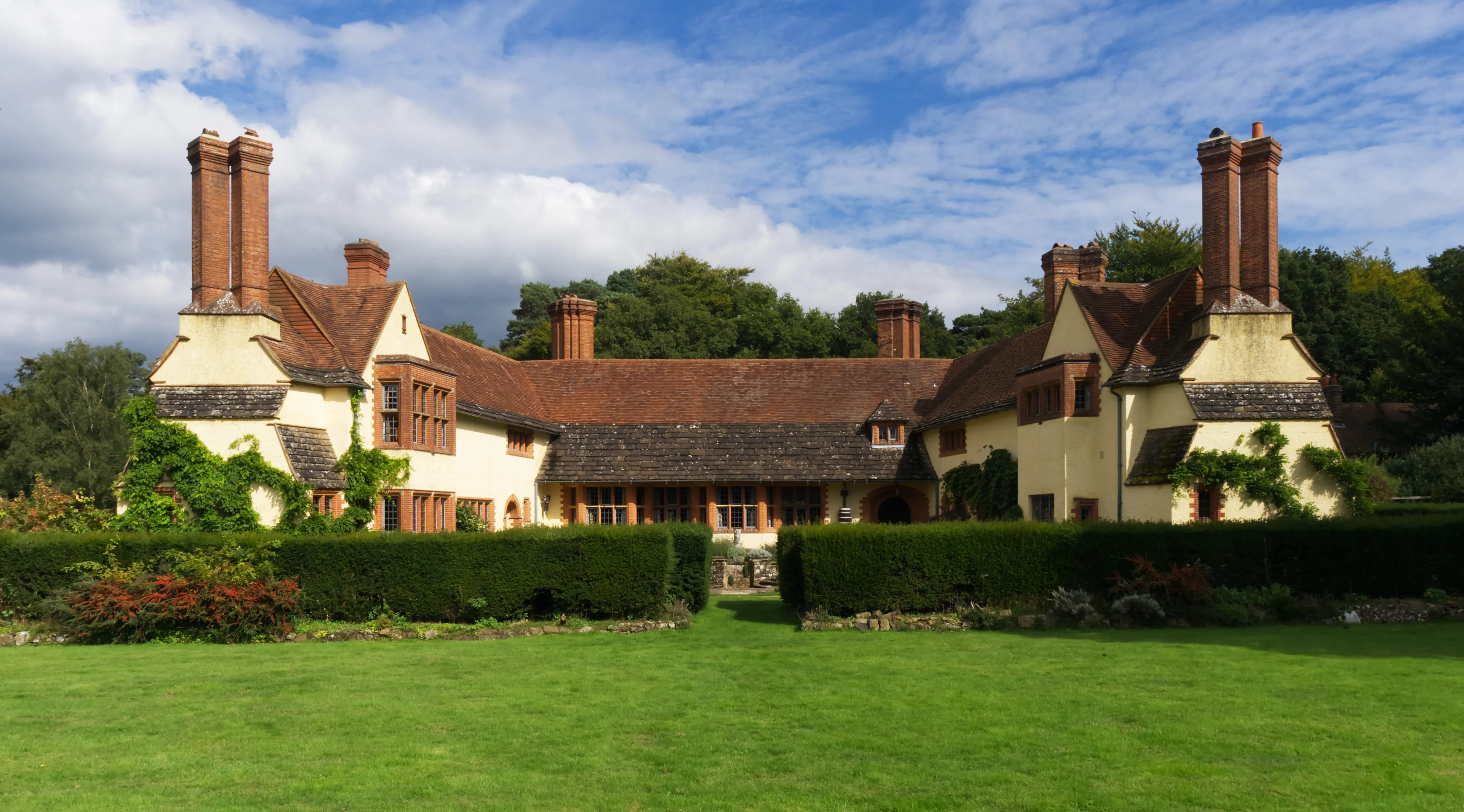 La casa de campo Goddards con sus altas chimeneas de ladrillo, tejado de tejas rojas y paredes color crema, vista a través de un césped verde con setos cuidados bajo un cielo azul