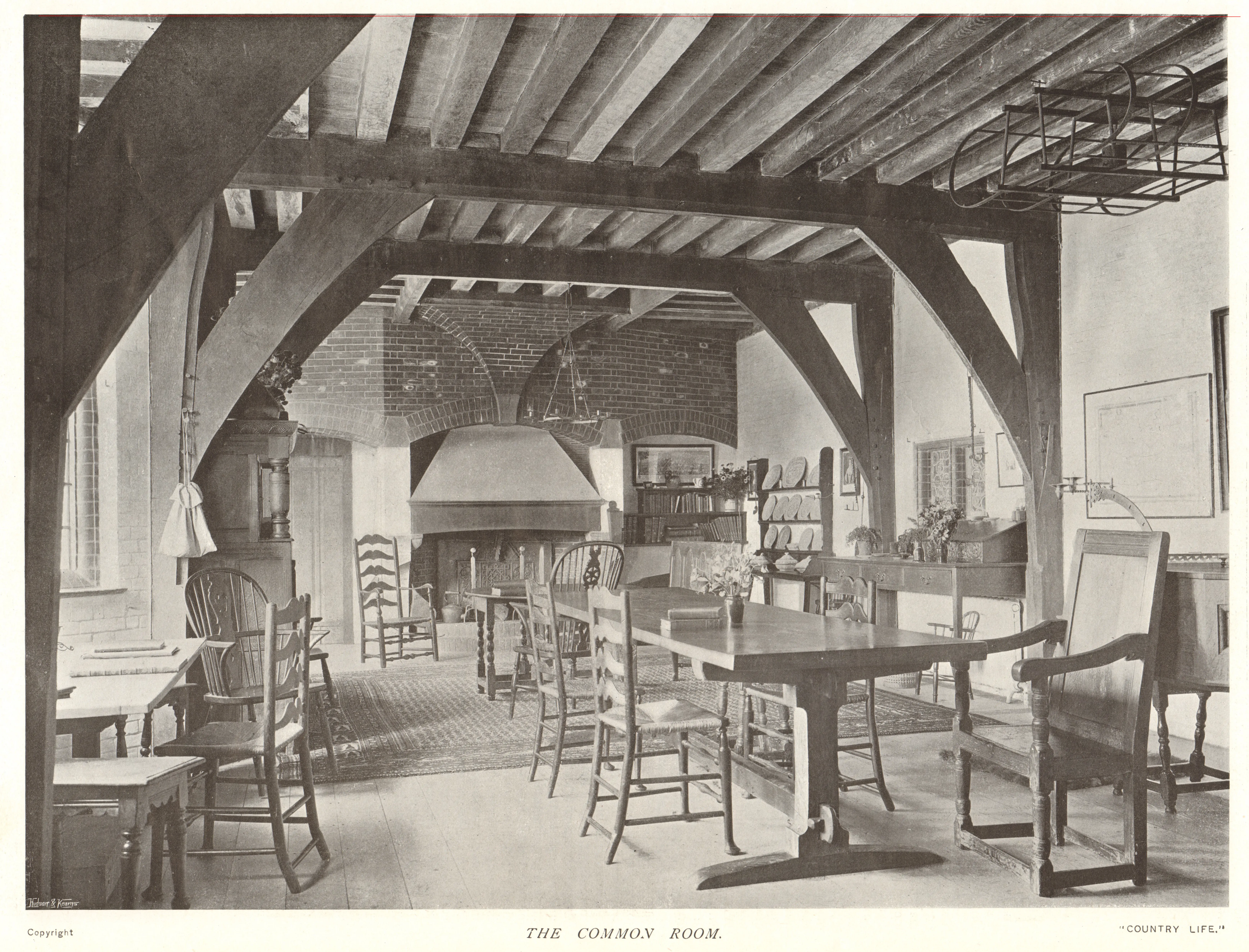 Fotografía en blanco y negro de la Common Room en Goddards, que muestra el techo con vigas de madera a la vista, una gran chimenea, mesas y sillas de madera, y estanterías.